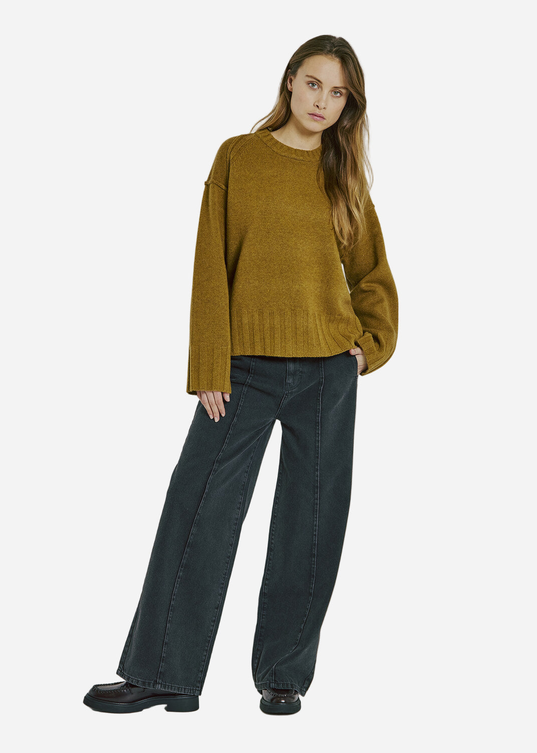 Woman wearing a dark mustard yellow sweater and dark jeans on a plain background