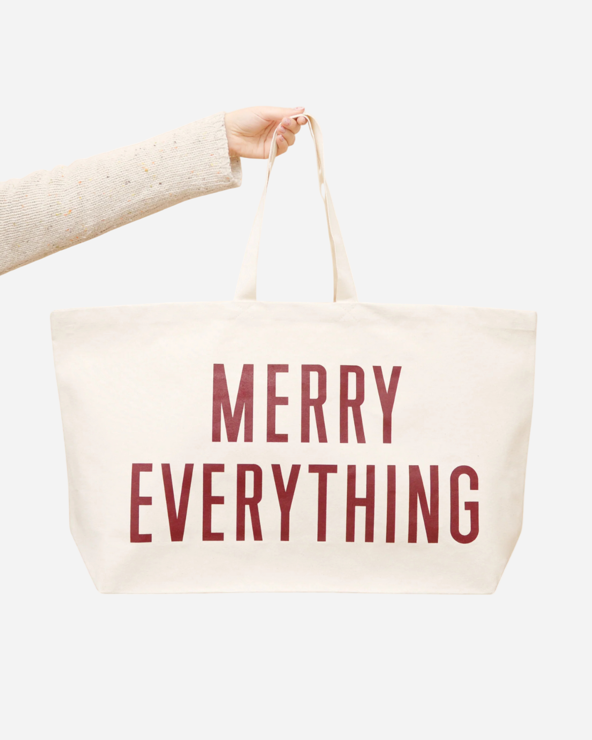 Beige tote bag with 'Merry Everything' text held by a person on a white background