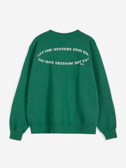 A green crewneck sweatshirt shown from the back, featuring white text that reads 'LET THE MYSTERY FIND YOU' in a circular design.