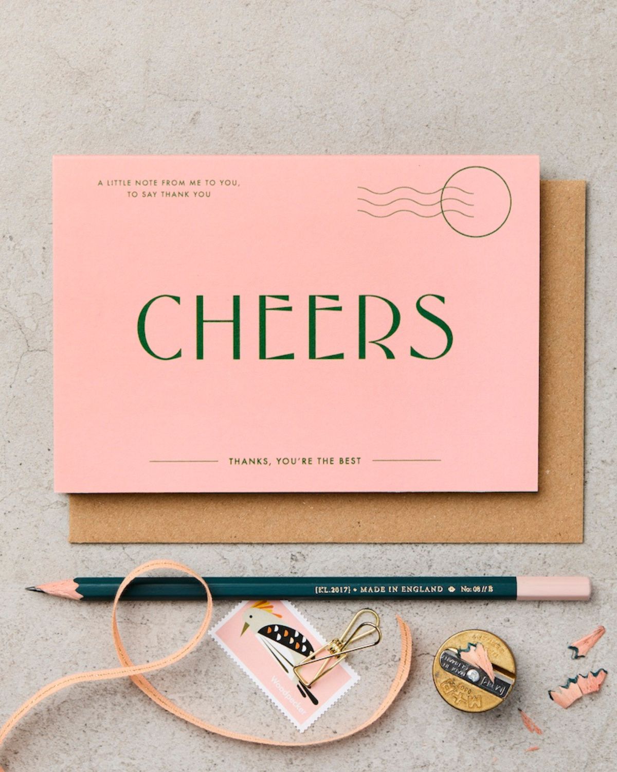 Pink card with 'Cheers' text on a textured surface with stationery items.