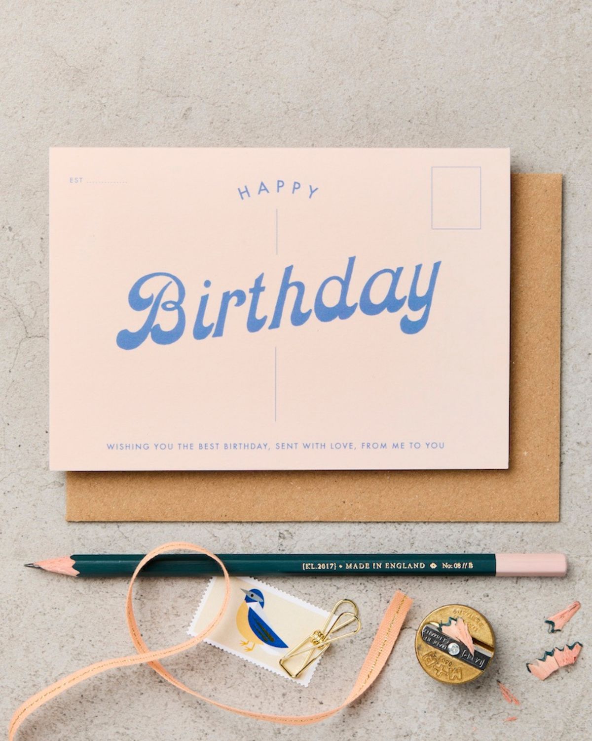 Birthday card with blue text on a beige background, accompanied by a pencil and ribbon.