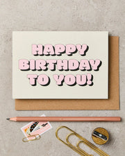 Birthday card with 'Happy Birthday to You!' text, pencil, paper clips, and stationery items on a textured surface.