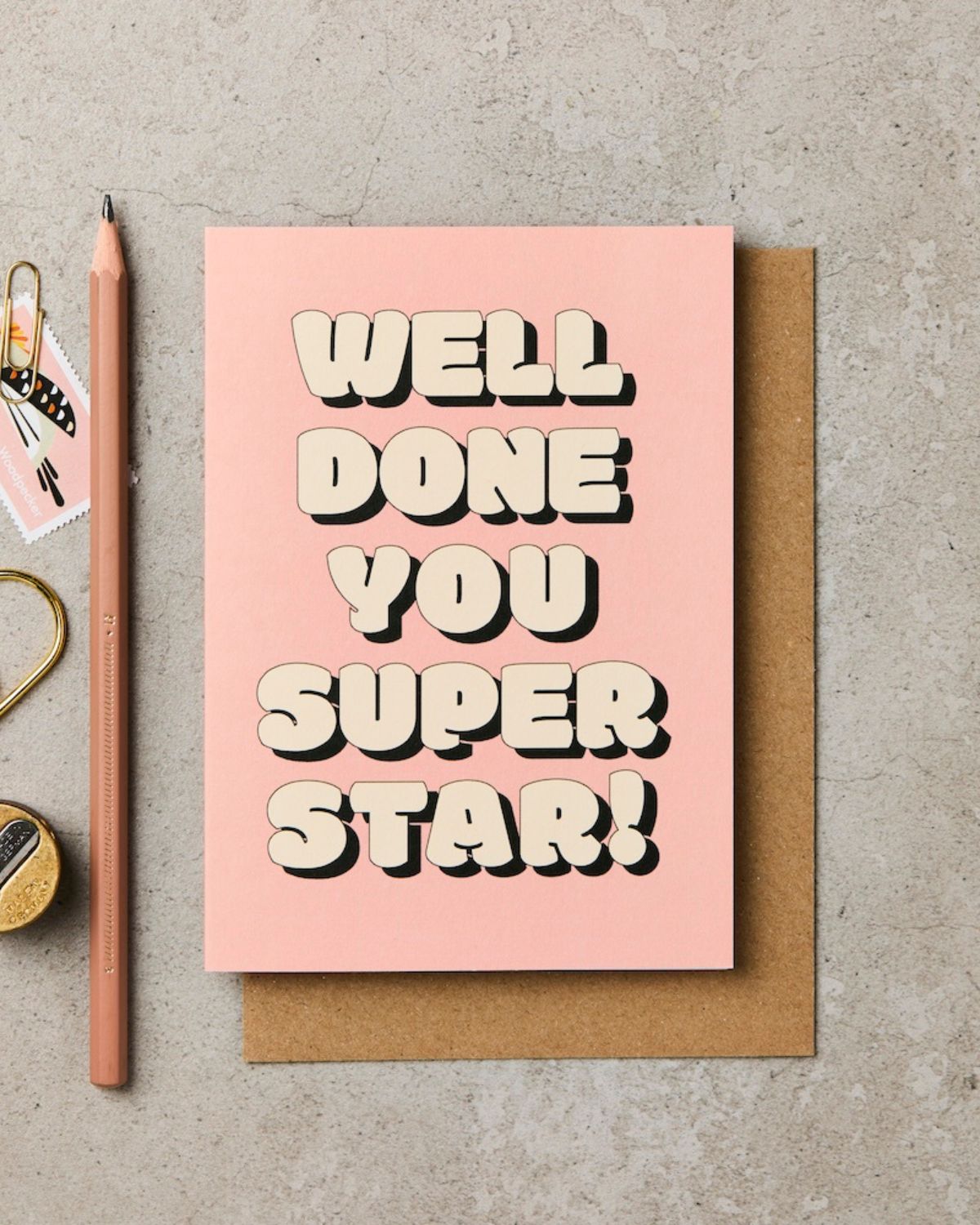 Pink card with 'Well done you super star!' text on a gray surface with a pencil and decorative items.