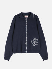 A dark navy blue knit cardigan sweater with white embroidered details, including a rose motif on the lower left chest and white bead trim along the collar and front button placket, shown against a plain light gray background.