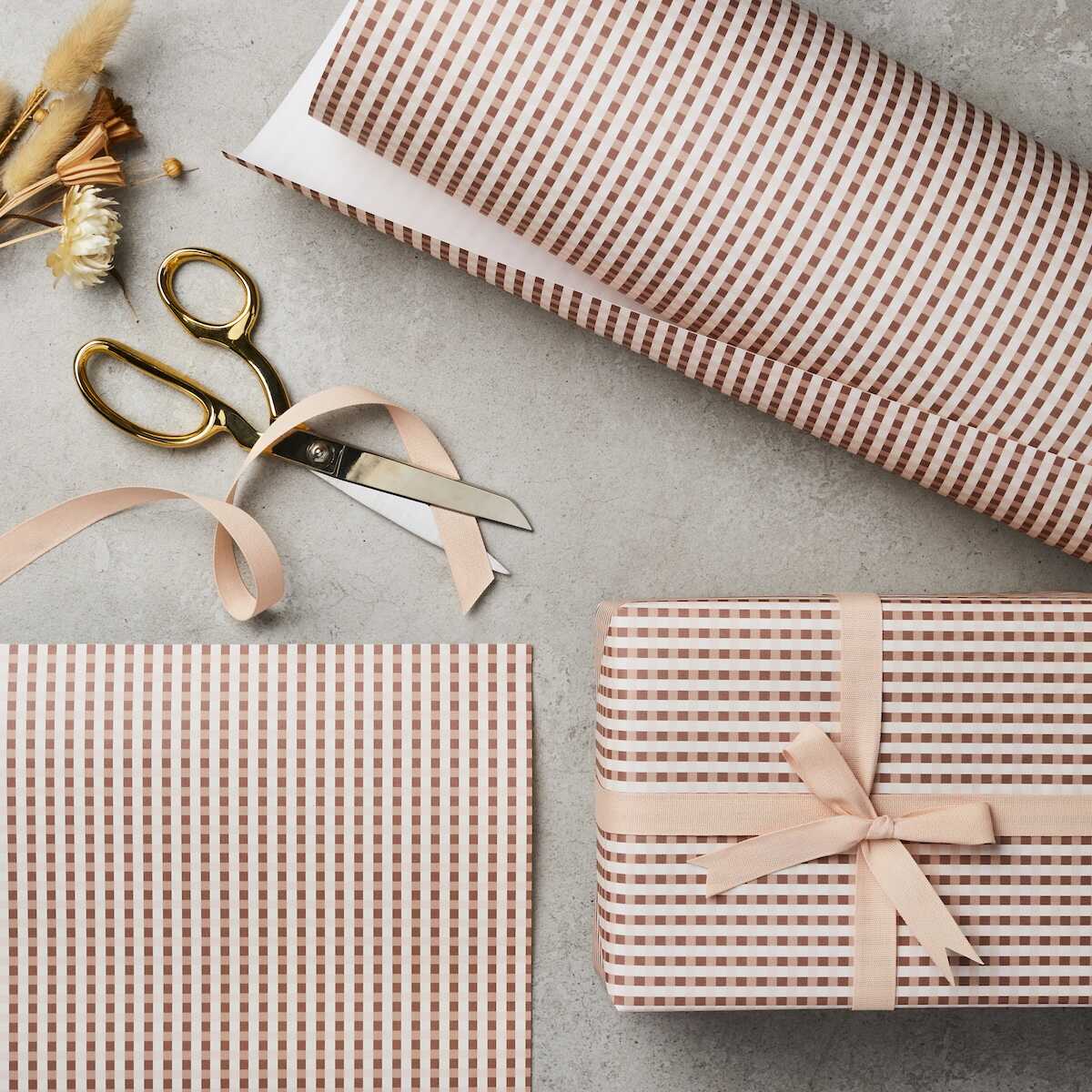 A roll of wrapping paper with a gingham pattern in a warm PINK shade, displayed next to a pair of scissors and a tied ribbon on a gift.
