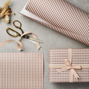 A roll of wrapping paper with a gingham pattern in a warm PINK shade, displayed next to a pair of scissors and a tied ribbon on a gift.