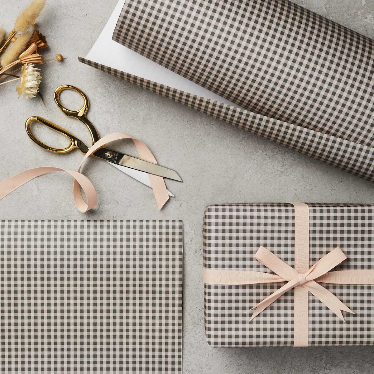 A roll of gingham wrapping paper in pink and green checkered pattern, alongside a pair of scissors and a tied ribbon on a gift box.