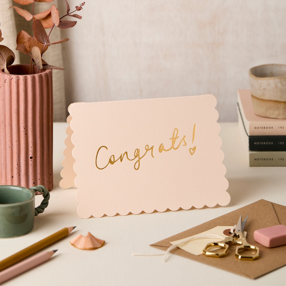 Pink card with 'Congrats!' text on a desk with stationery items