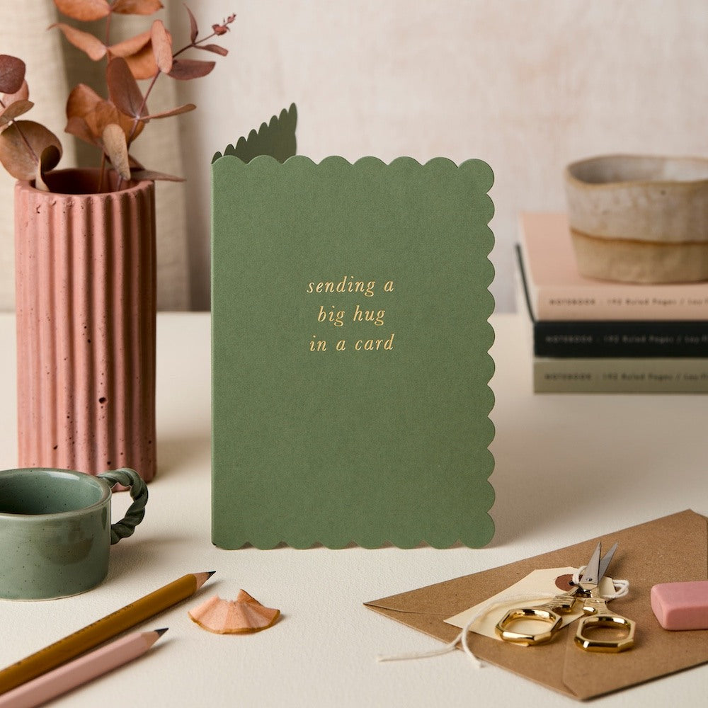 Green card with 'sending a big hug in a card' text on a desk with stationery items.