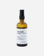 A glass bottle of Meraki room spray with a black spray nozzle. The label shows the product name and the scent, Rain Forest.