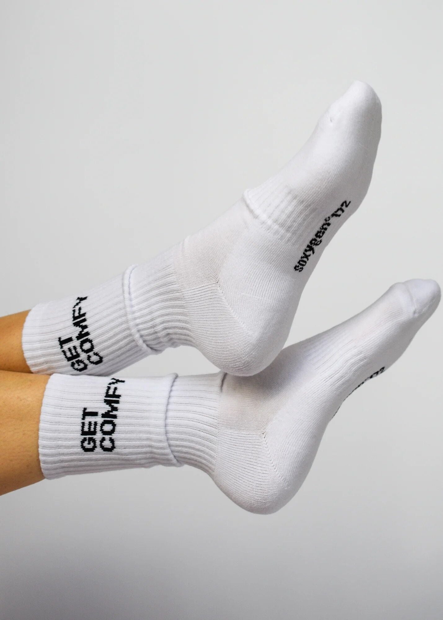 A pair of white crew-length socks with the text 'GET COMFY' printed on the leg, featuring arch support and cushioned heel and toe areas.
