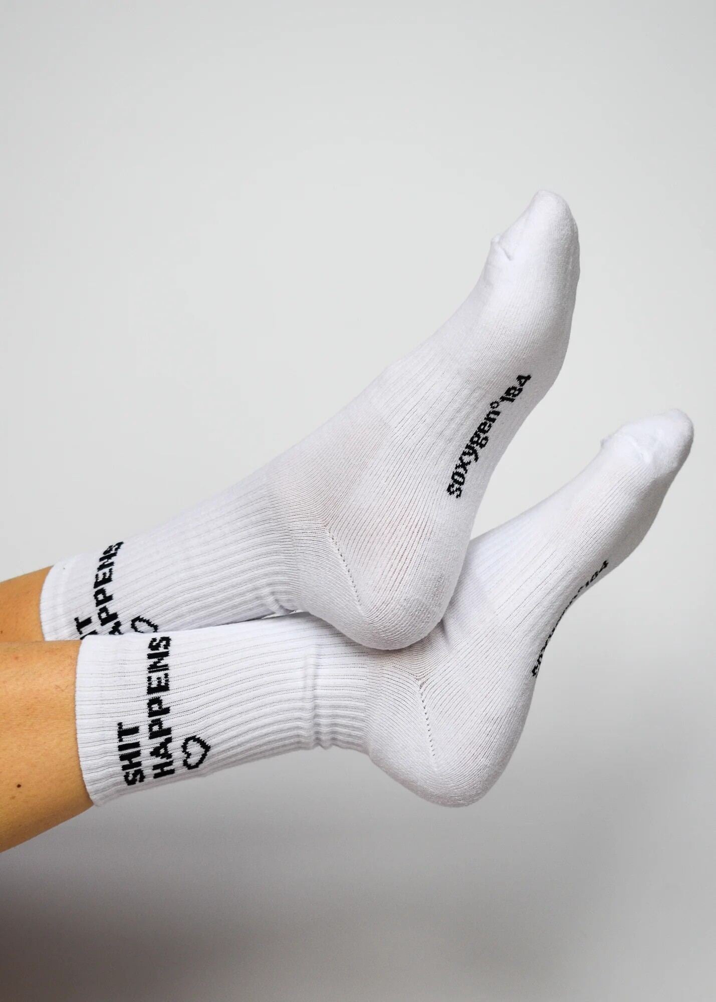 A pair of white crew-length socks with the phrase 'SHIT HAPPENS' written on the side, featuring arch support and cushioned heel and toe areas.
