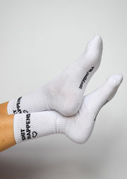 A pair of white crew-length socks with the phrase 'SHIT HAPPENS' written on the side, featuring arch support and cushioned heel and toe areas.