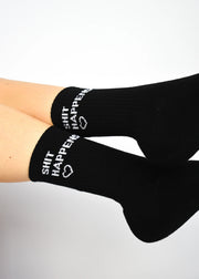A pair of black crew length socks with the slogan 'SHIT HAPPENS' written on the side, worn on a person's legs.