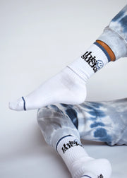 A pair of white crew-length socks with the text 'THIS SUCKS' and a smiley face design in black, placed on a person's legs against a tie-dye background.