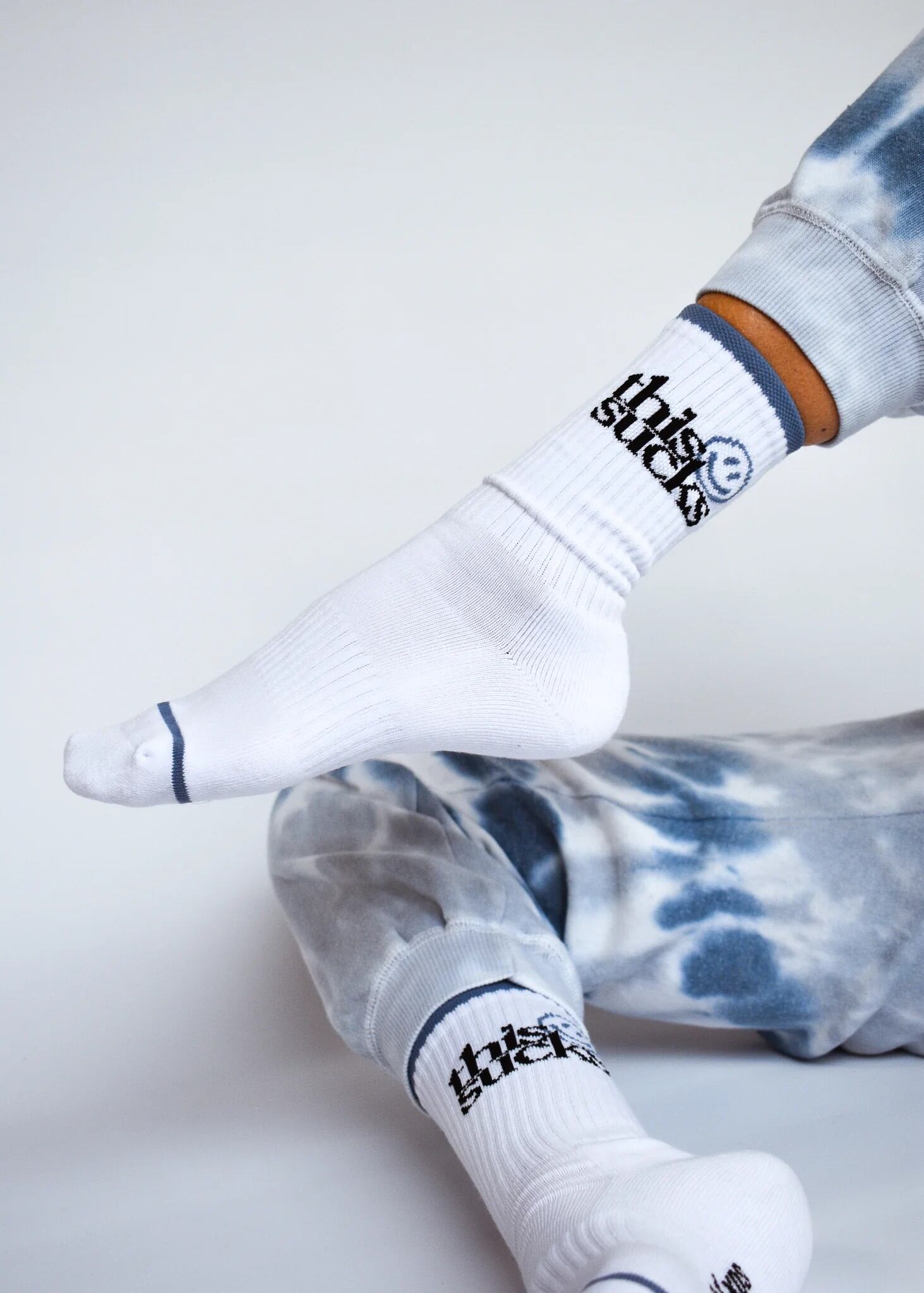 A pair of white crew-length socks with the text 'THIS SUCKS' and a smiley face design in black, placed on a person's legs against a tie-dye background.