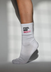A pair of white crew length socks with 'FEEL GOOD' ombre slogan detail, showing arch support and cushioned heel and toe.