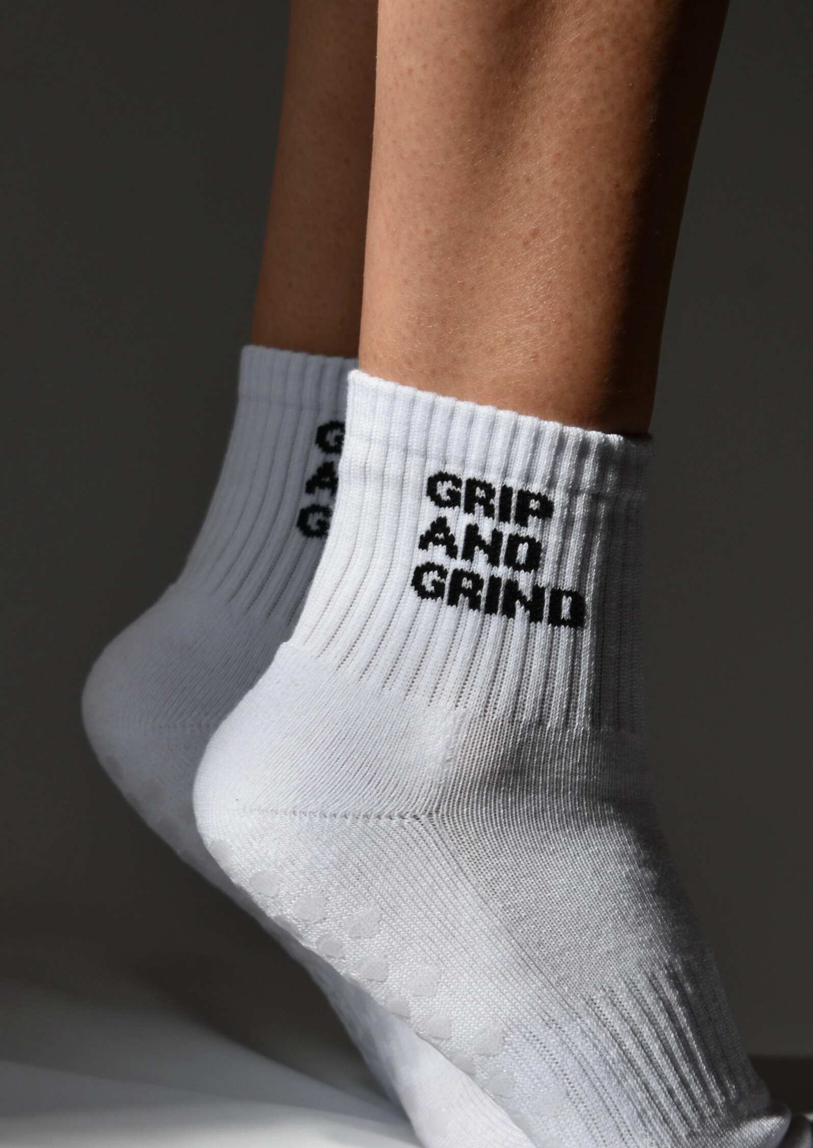A pair of white crew-length socks with 'GRIP AND GRIND' text on the side, featuring a non-slip sole design.