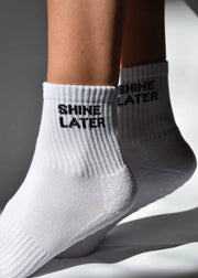 A pair of white grip socks with 'SHINE LATER' printed on the side, positioned on a person's feet against a white background.