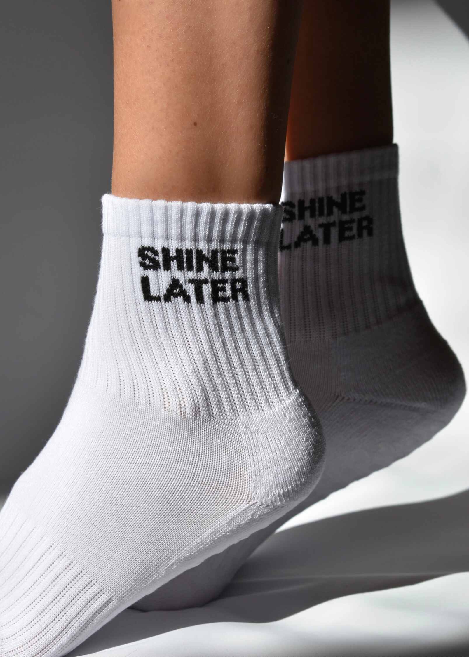 A pair of white grip socks with 'SHINE LATER' printed on the side, positioned on a person's feet against a white background.