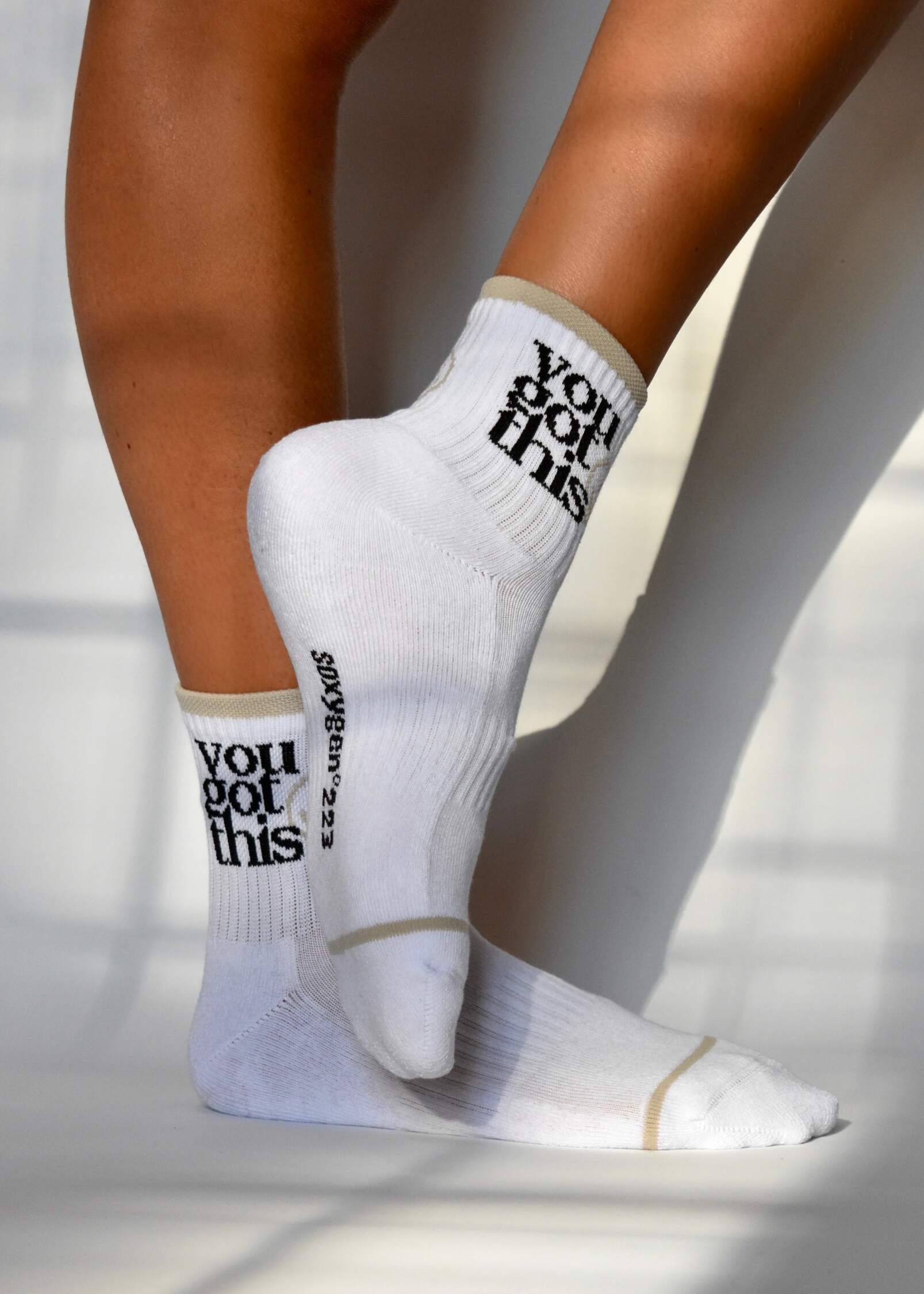 A pair of white ribbed crew socks with the text 'YOU GOT THIS - SMILE' printed on the side, worn on legs raised in the air against a white background.