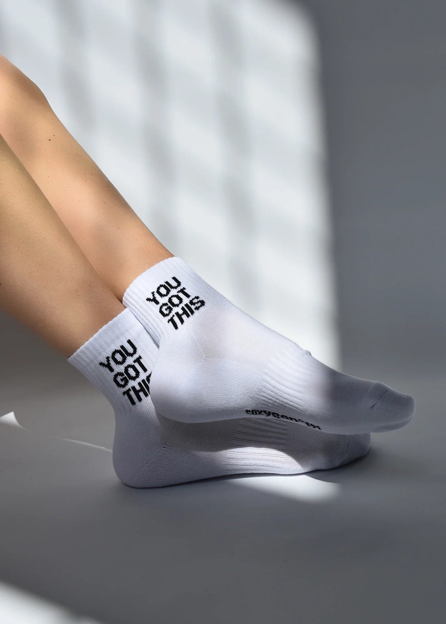 A pair of white ribbed crew socks with 'YOU GOT THIS' text detail, worn on a person's ankles.
