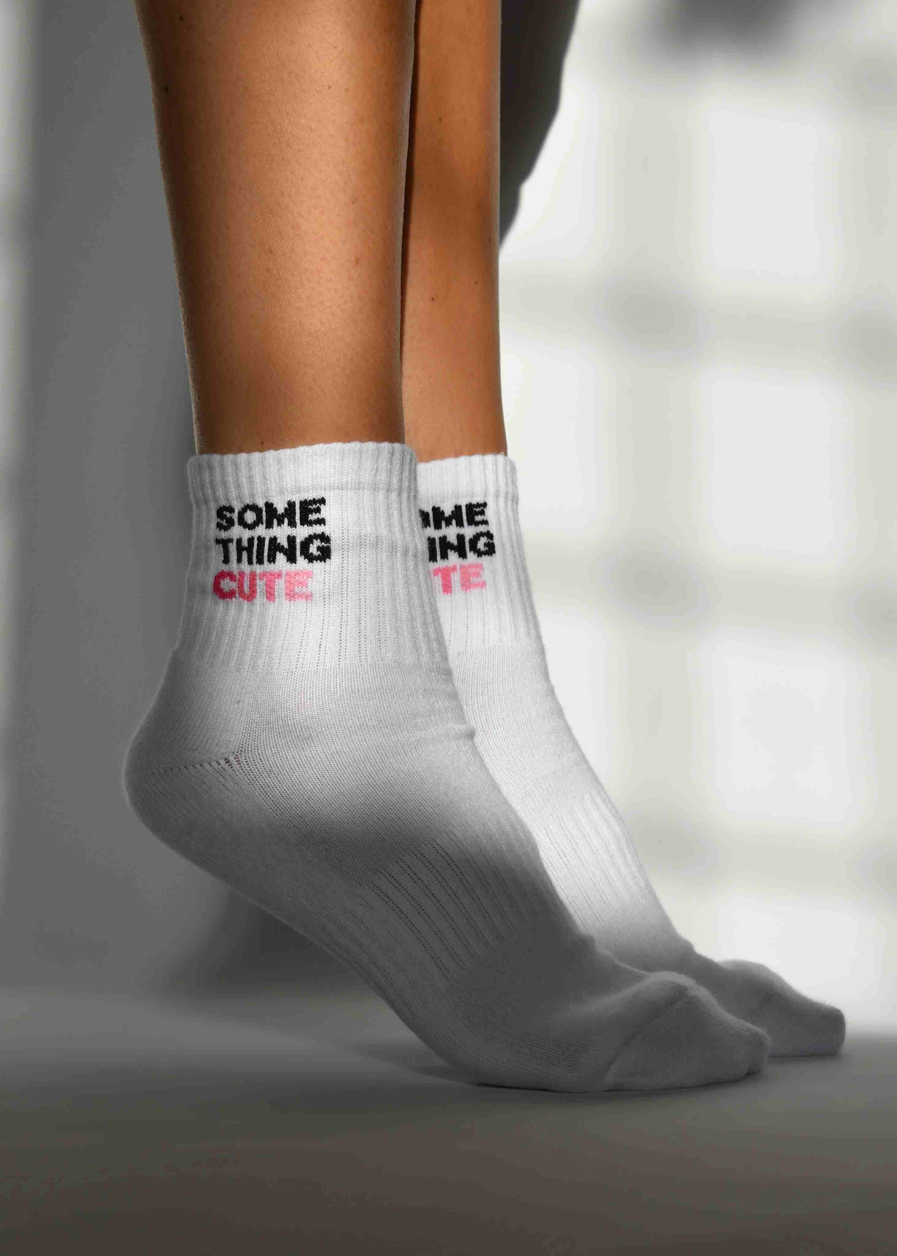 A pair of white ribbed crew socks with 'SOMETHING CUTE' text detail, worn on the lower leg above the ankle.