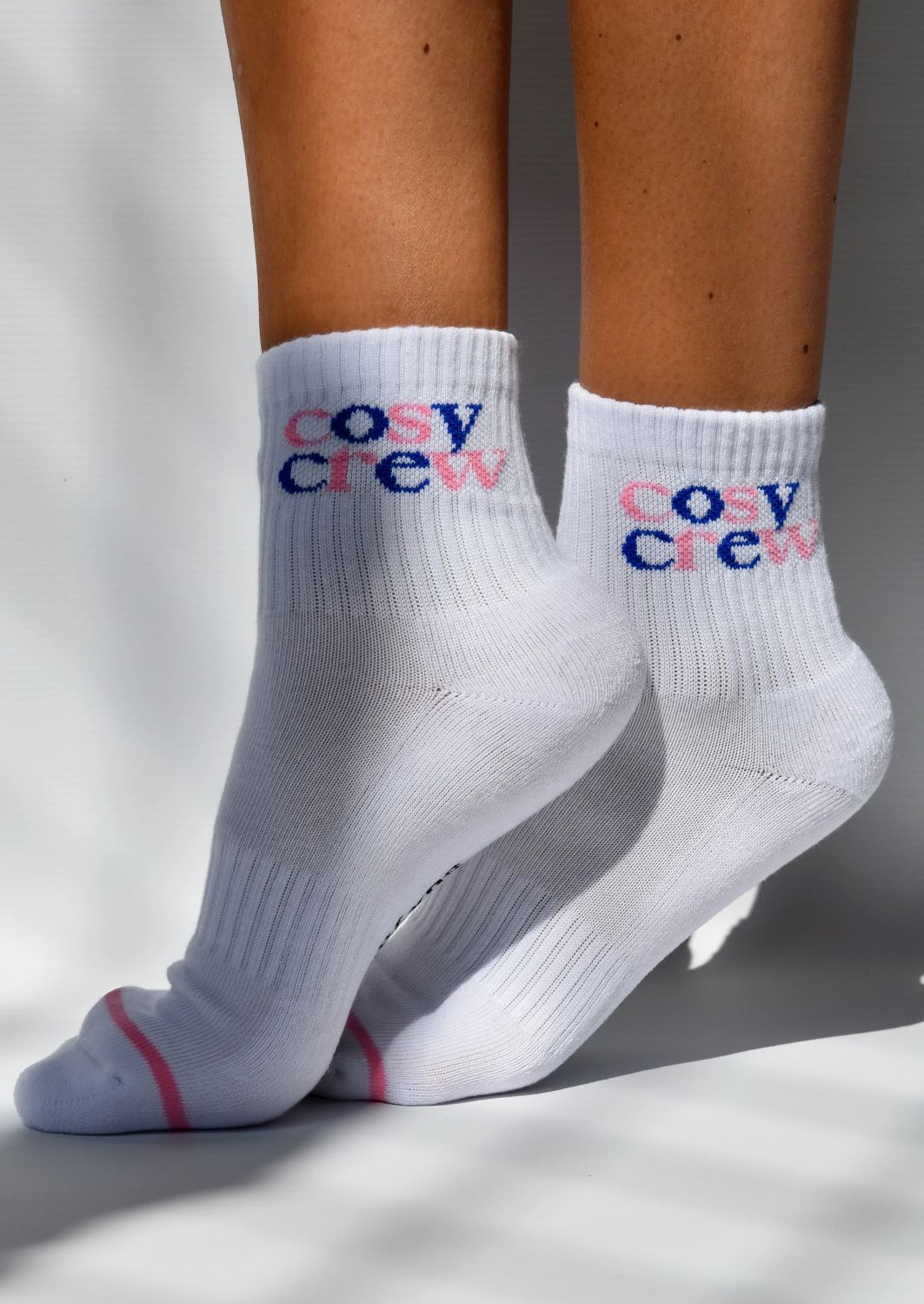 A pair of white ribbed crew socks with 'COSY CREW' text in red and blue details above the ankle.