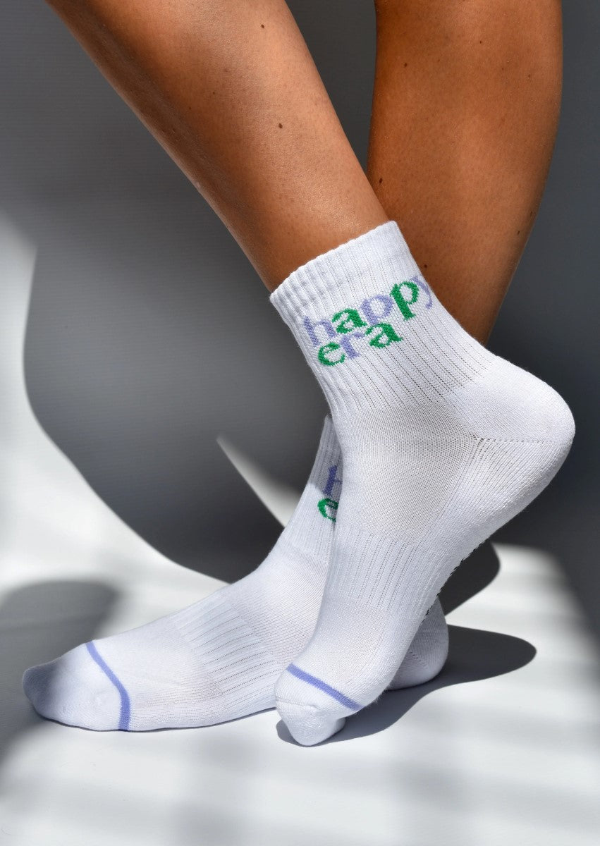A pair of white ribbed crew socks with 'HAPPY ERA' text in green detail, showing the socks worn on feet against a light background.